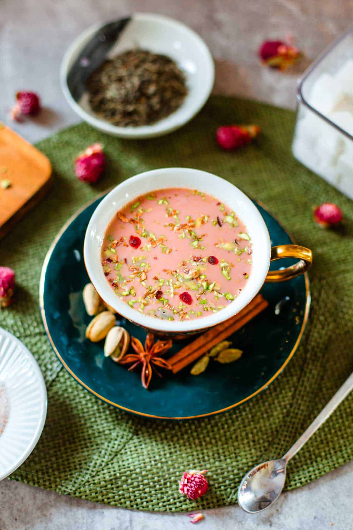 kashmiri pink chai in teacup surrounded by flowers, tea leaves, sugar cubes, spices.