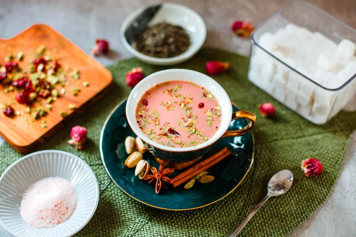 kashmiri pink chai in teacup surrounded by flowers, tea leaves, sugar cubes, spices.