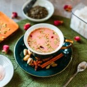 kashmiri pink chai in teacup surrounded by flowers, tea leaves, sugar cubes, spices.