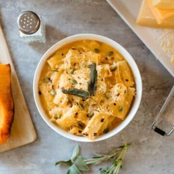 copycat trader joe's butternut squash mac and cheese in bowl surrounded by herbs, squash, cheese blocks, grater, salt shaker.
