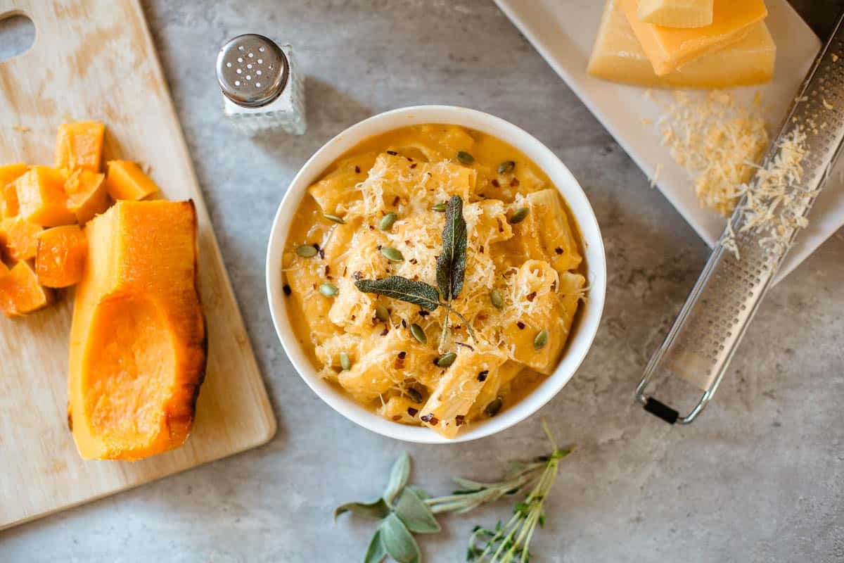 copycat trader joe's butternut squash mac and cheese in bowl surrounded by herbs, squash, cheese blocks, grater, salt shaker.