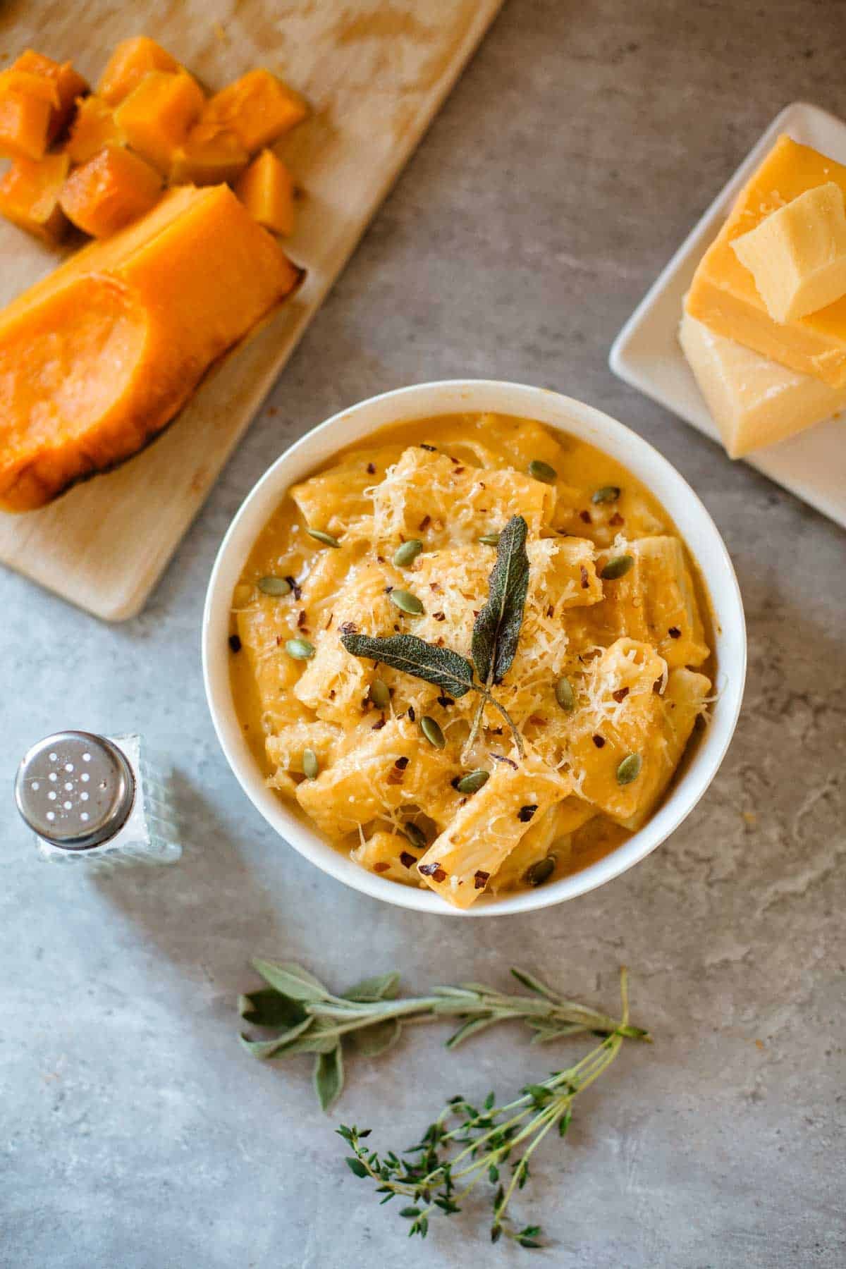 copycat trader joe's butternut squash mac and cheese in bowl surrounded by herbs, squash, cheese blocks, grater, salt shaker.