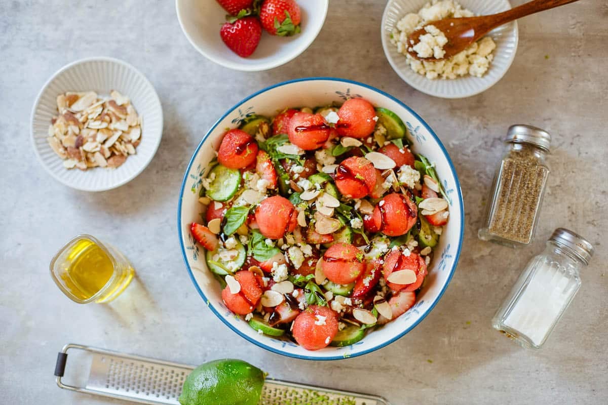 watermelon salad in a bowl surrounded by toasted almonds, strawberries, lime, cheese, seasonings.