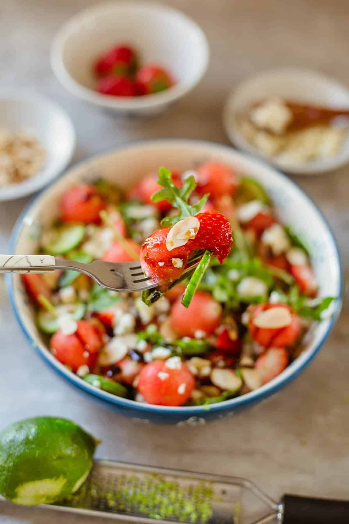 fruits and veggies on a fork above the bowl of watermelon salad and smaller bowls with toppinhs (nuts, fruit, cheese, etc.)