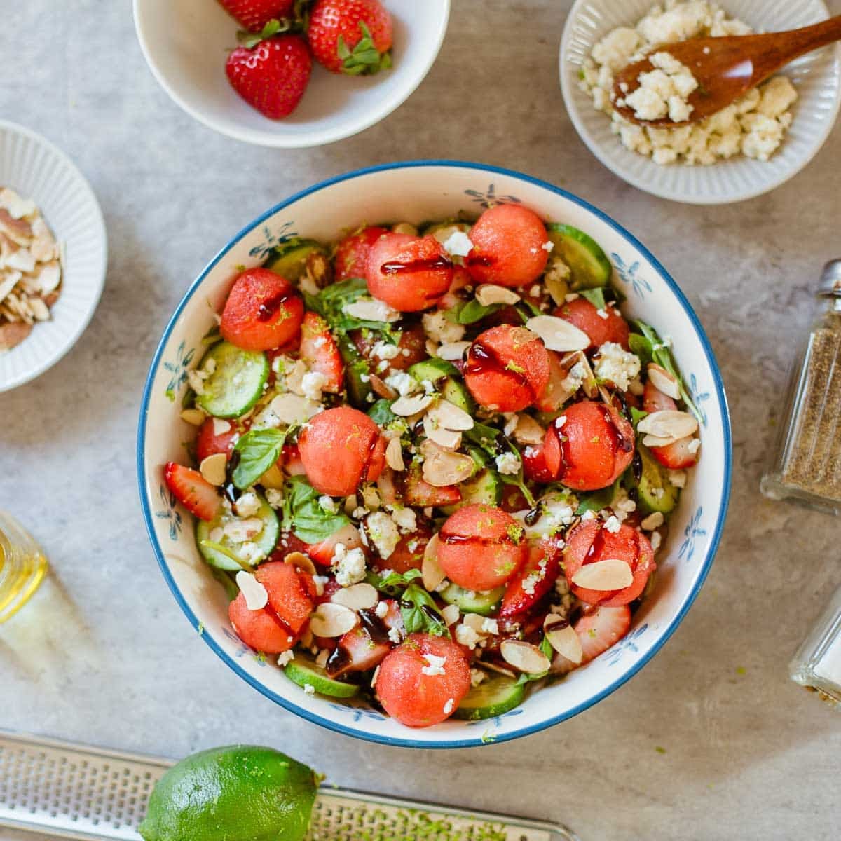 watermelon salad in a bowl surrounded by toasted almonds, strawberries, lime, cheese, seasonings.