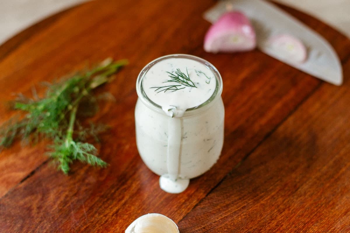 dill dressing dripping from jar beside dill, garlic, shallot, and knife.