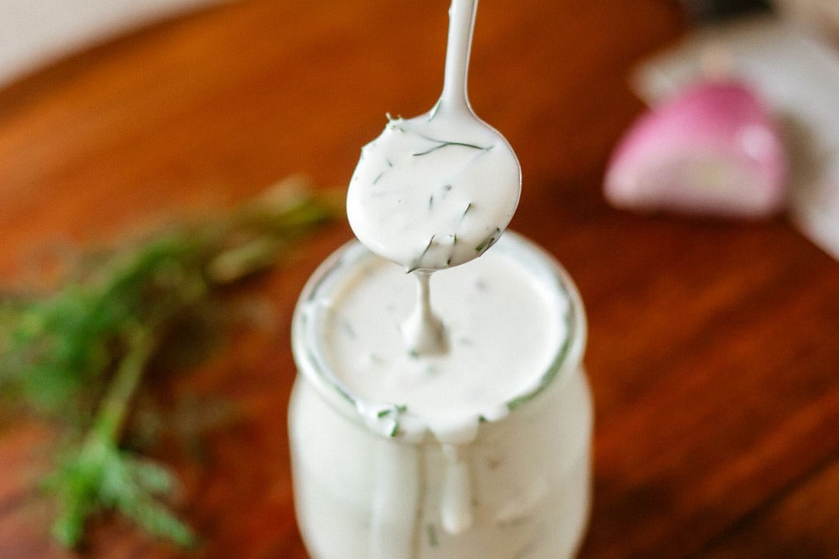 spoon drizzling dill dressing from jar beside shallot and herbs.