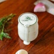 dressing dripping from jar beside dill, garlic, shallot, and knife.