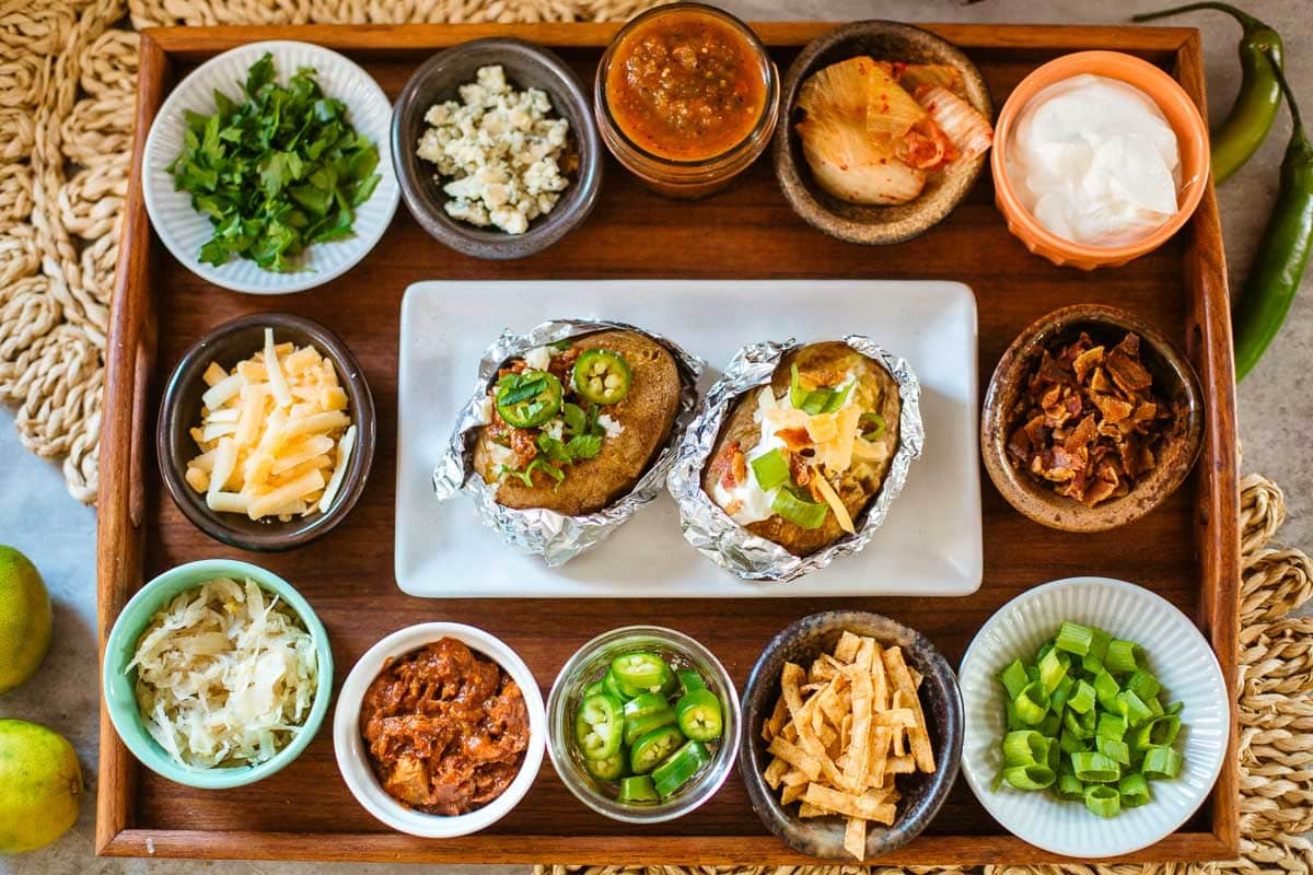 various baked potato bar toppings (cheese, meat, veggies, etc.) in small bowls with baked potatoes.