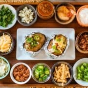 various baked potato bar toppings (cheese, meat, veggies, etc.) in small bowls with baked potatoes.