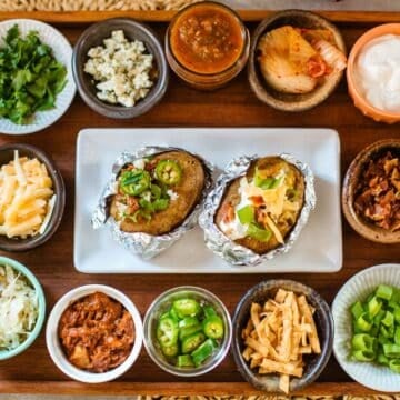 various baked potato bar toppings (cheese, meat, veggies, etc.) in small bowls with baked potatoes.