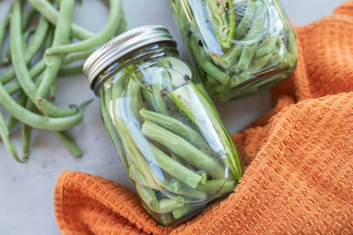 2 jars of pickled green beans with green beans and an orange towel. 