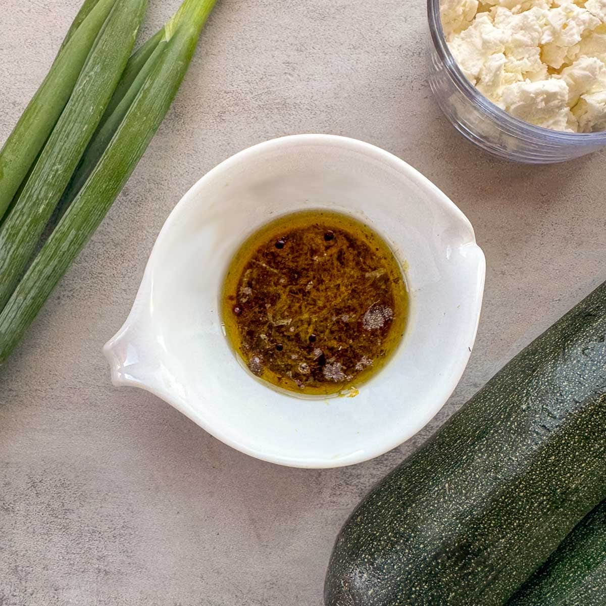oil dressing in a small bowl with feta, zucchini, and green oinions around it. 