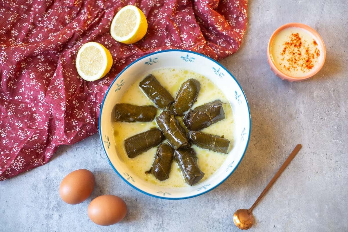 dolmades in avgolemono sauce beside lemons, eggs, spoon, bowl of sauce.