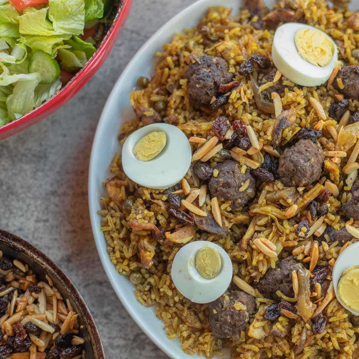 chicken biryani with a bowl of salad and some fried raisins and almonds next to it.