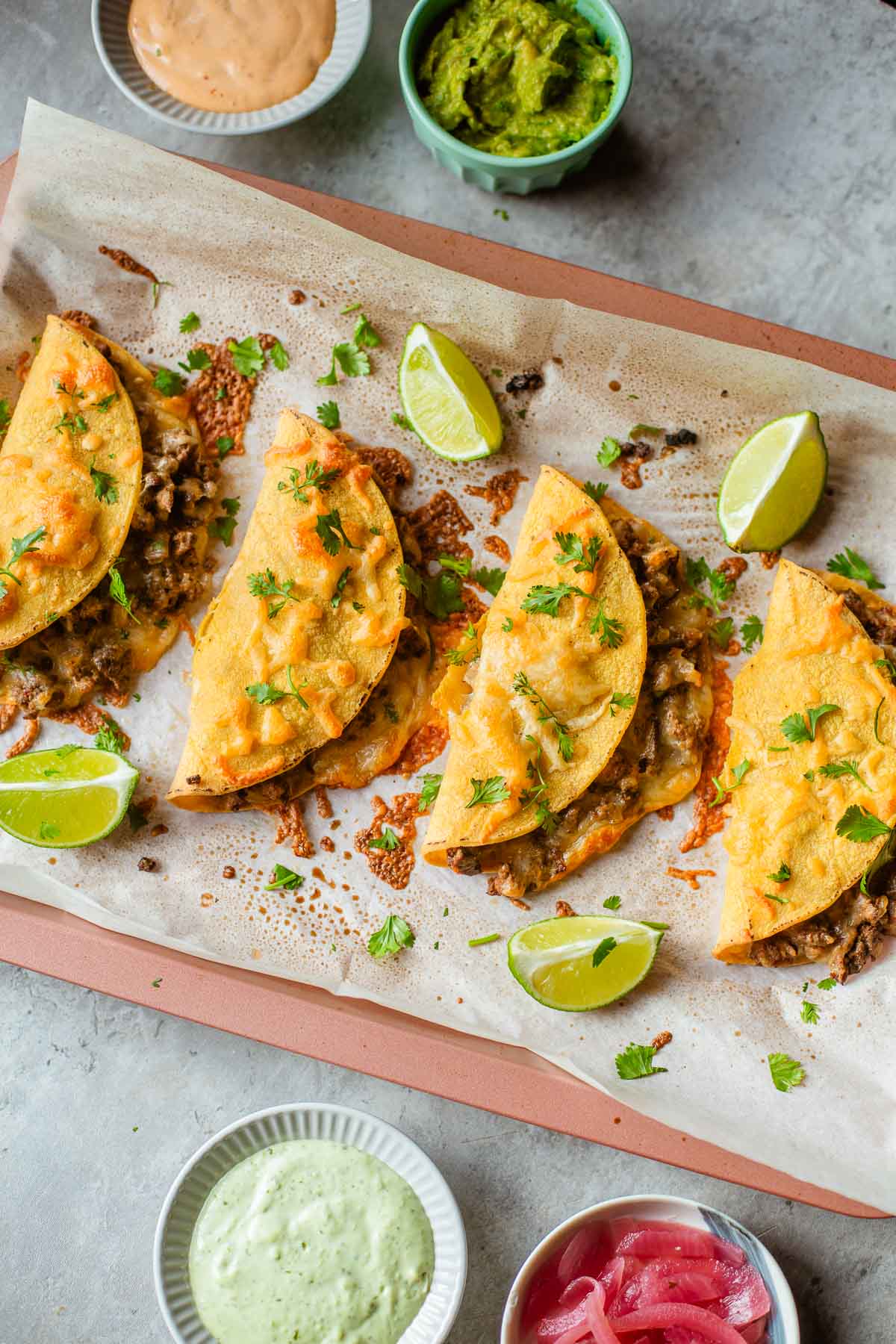sheet pan tacos topped with cilantro beside lime wedges and various bowls of sauces and toppings (guacamole, pickled onions, etc.)