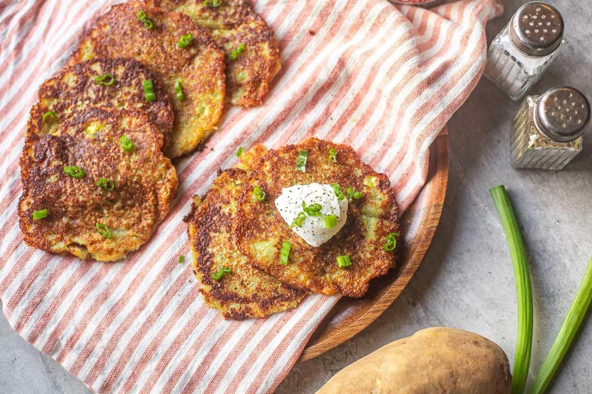 Deruny on striped mat topped with sour cream and green onions beside onion, potato, salt and pepper.