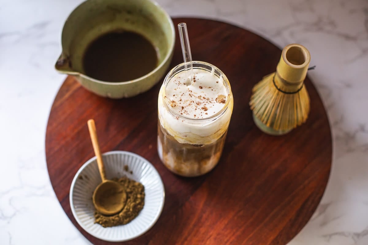 hojicha latte on wooden board with hojicha powder, whisk, bowl of tea.