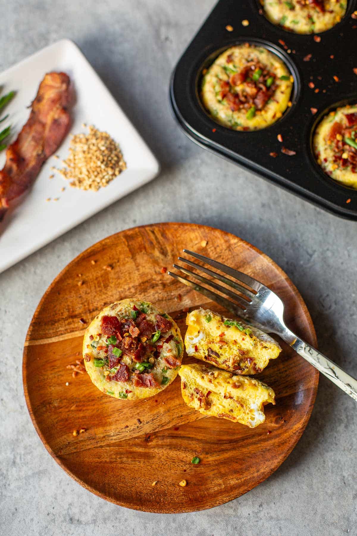 baked eggs in a muffin tin beside plates with halved egg, bacon, chives, sesame seeds.