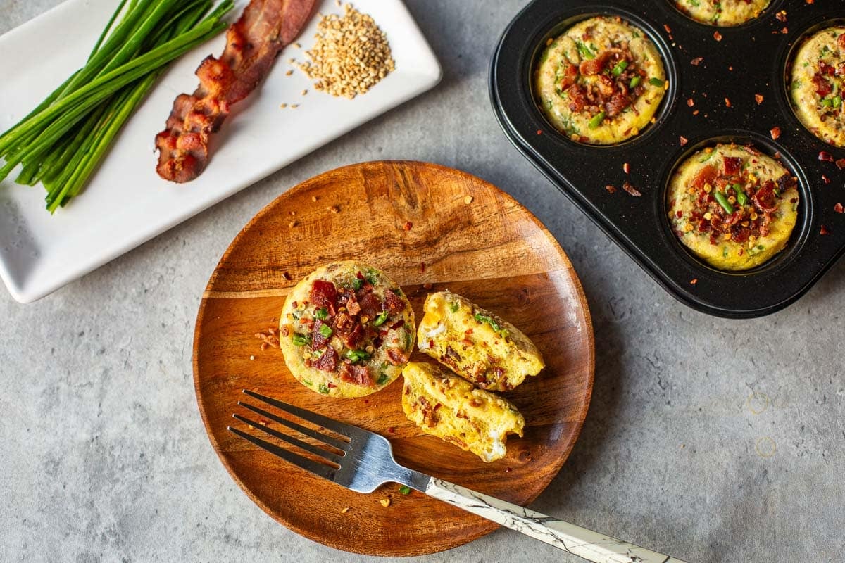 baked eggs in a muffin tin beside plates with halved egg, bacon, chives, sesame seeds.