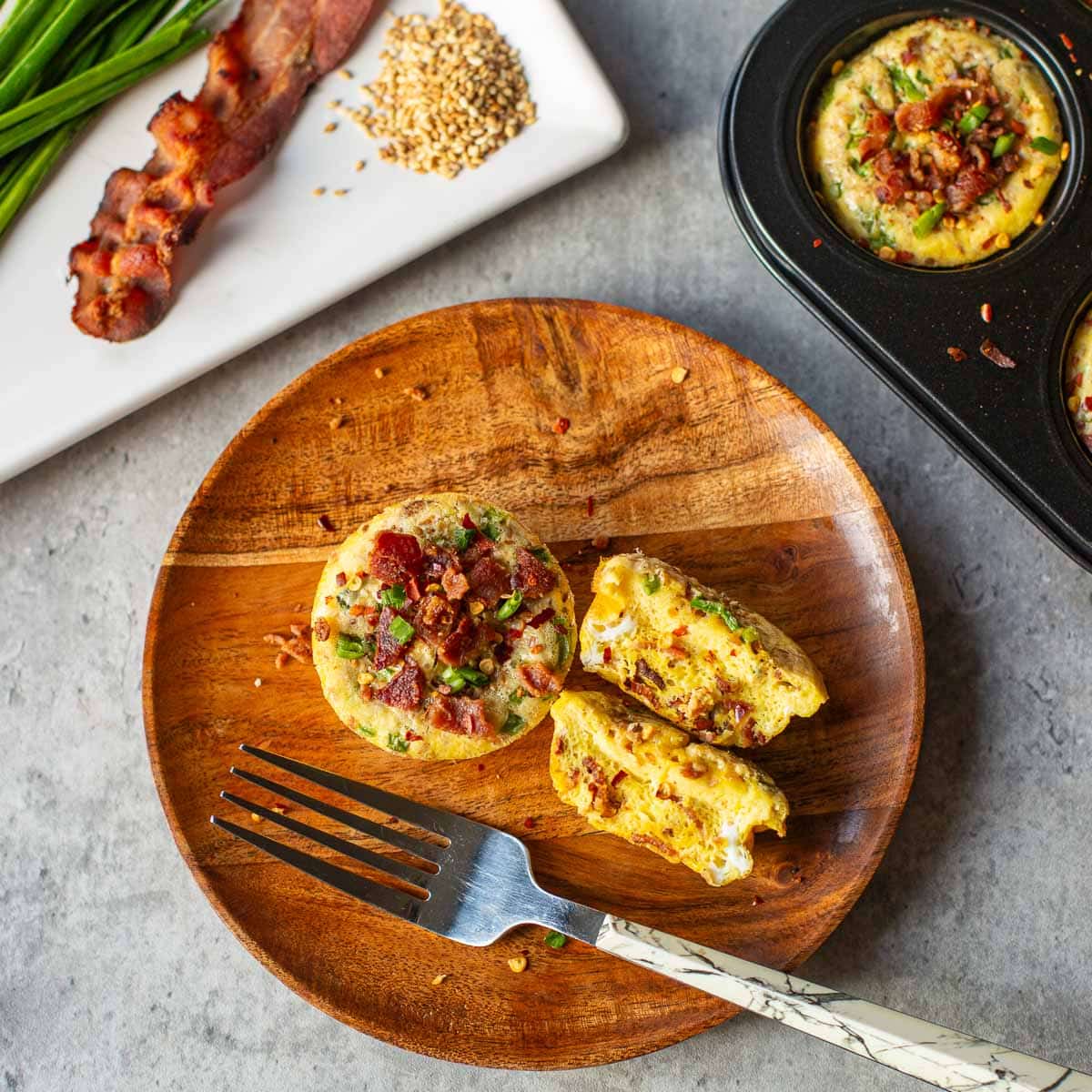 baked eggs in a muffin tin beside plates with halved egg, bacon, chives, sesame seeds.