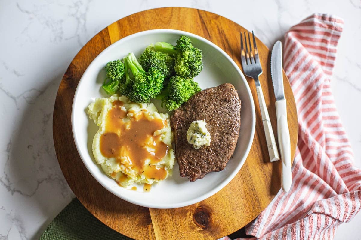 air fryer sirloin steak topped with herb butter beside broccoli, mashed potatoes, and utensils.
