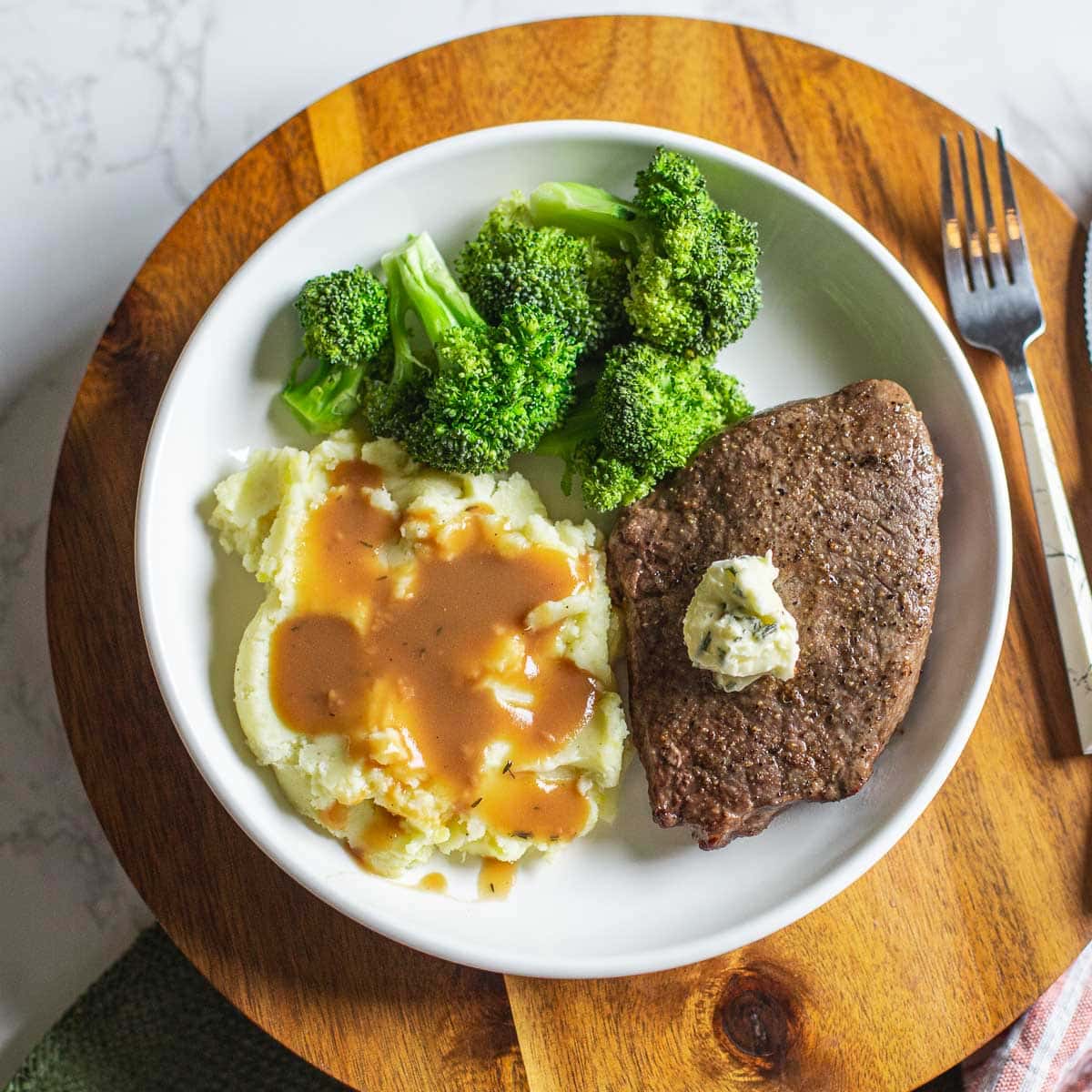 air fryer sirloin steak topped with herb butter beside broccoli, mashed potatoes, and utensils.
