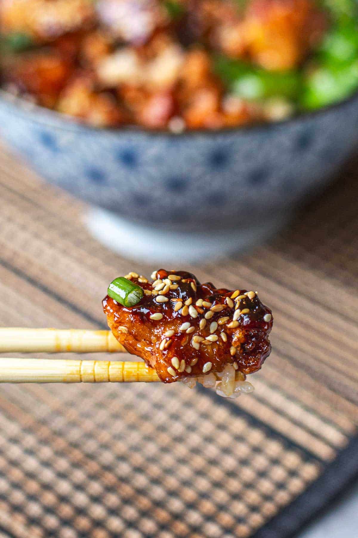 chopsticks holding saucy piece of sticky sesame chicken in front of bowl.