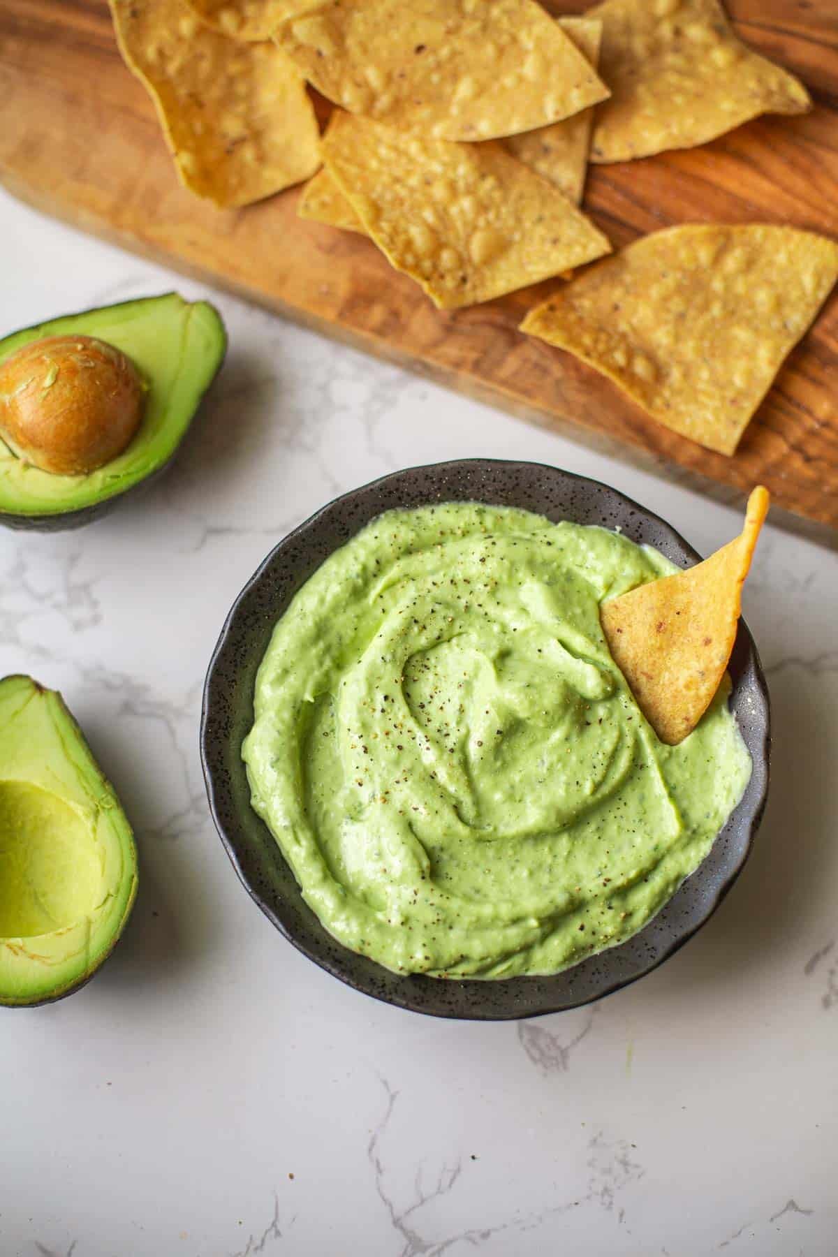 avocado sauce with chip dipped beside halved avocados and cutting board with lime & chips.