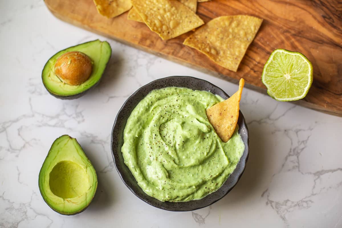 avocado sauce with chip dipped beside halved avocados and cutting board with lime & chips.