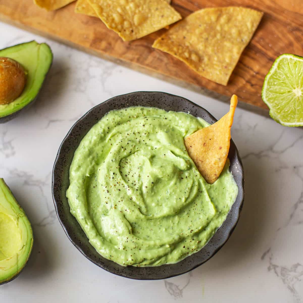avocado sauce with chip dipped beside halved avocados and cutting board with lime & chips.