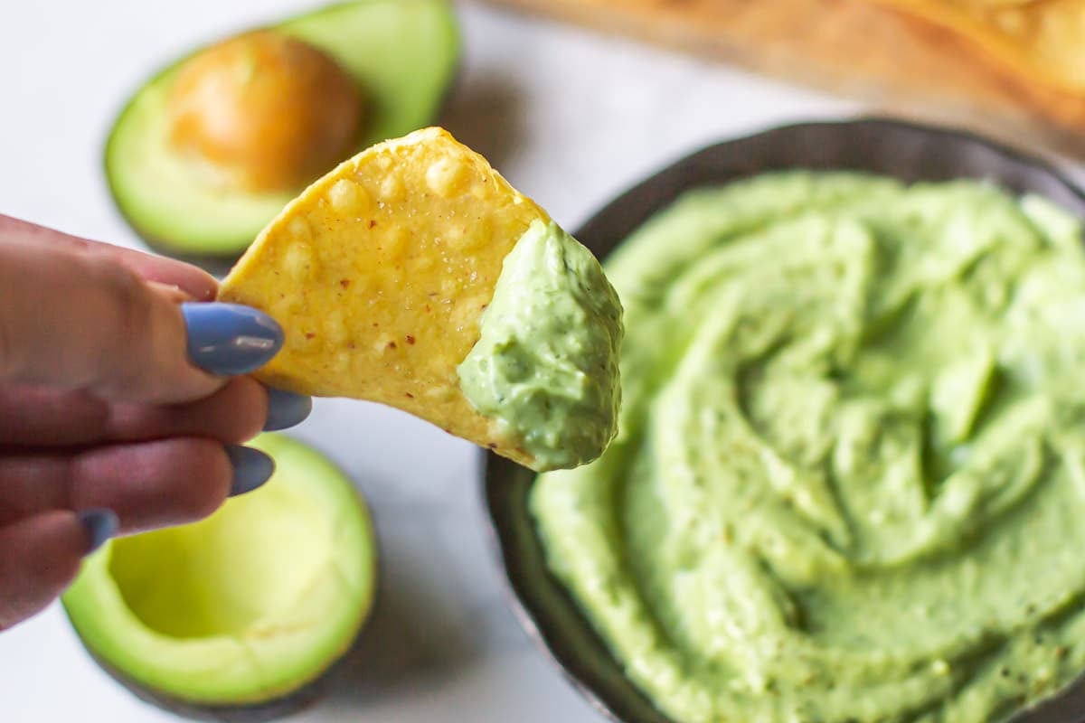 hand holding tortilla chip coated in avocado sauce above bowl and halved avocados.