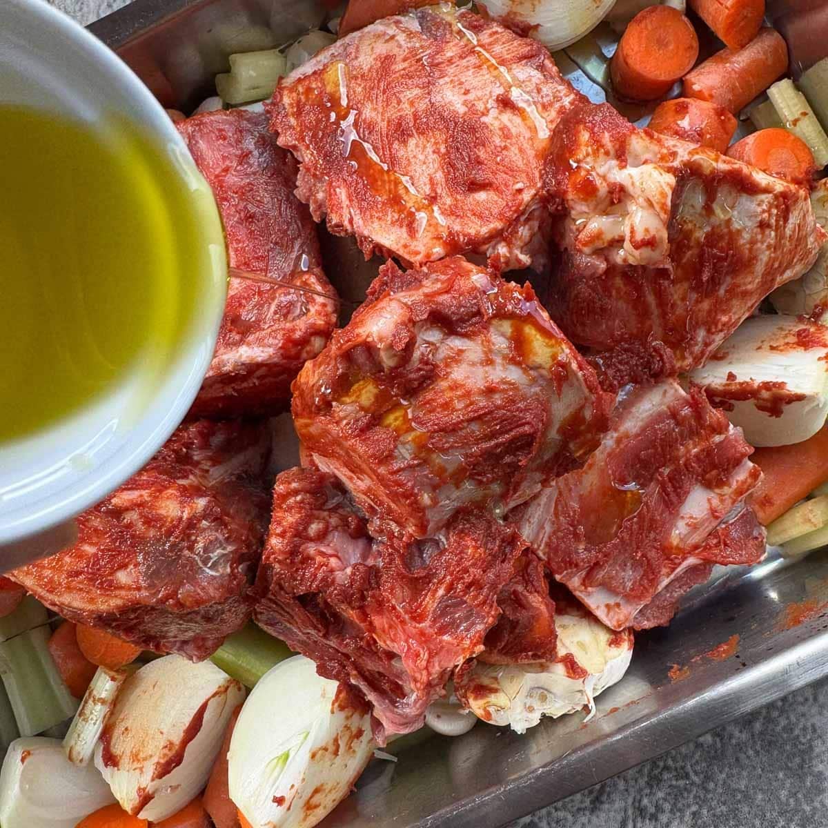 pouring olive oil over bones and veggies in a roasting pan for making beef bone broth