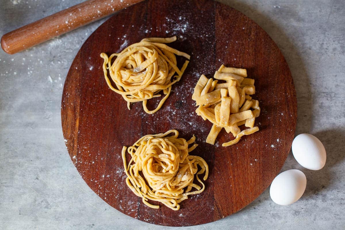 homemade egg noodles cut into three shapes: short & thick noodles, medium width noodles, and thin, delicate noodles beside rolling pin and eggs.