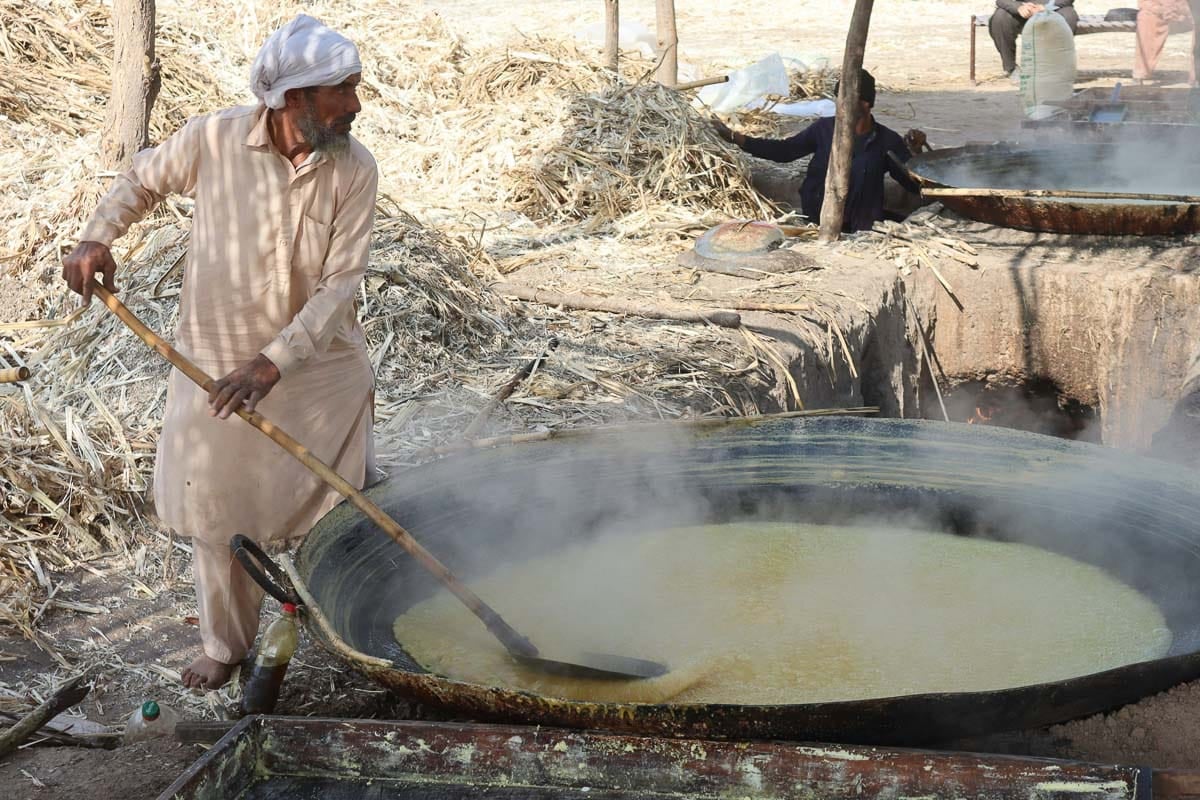 Indian man stirring giant pot of hot melted sugar in rural setting.