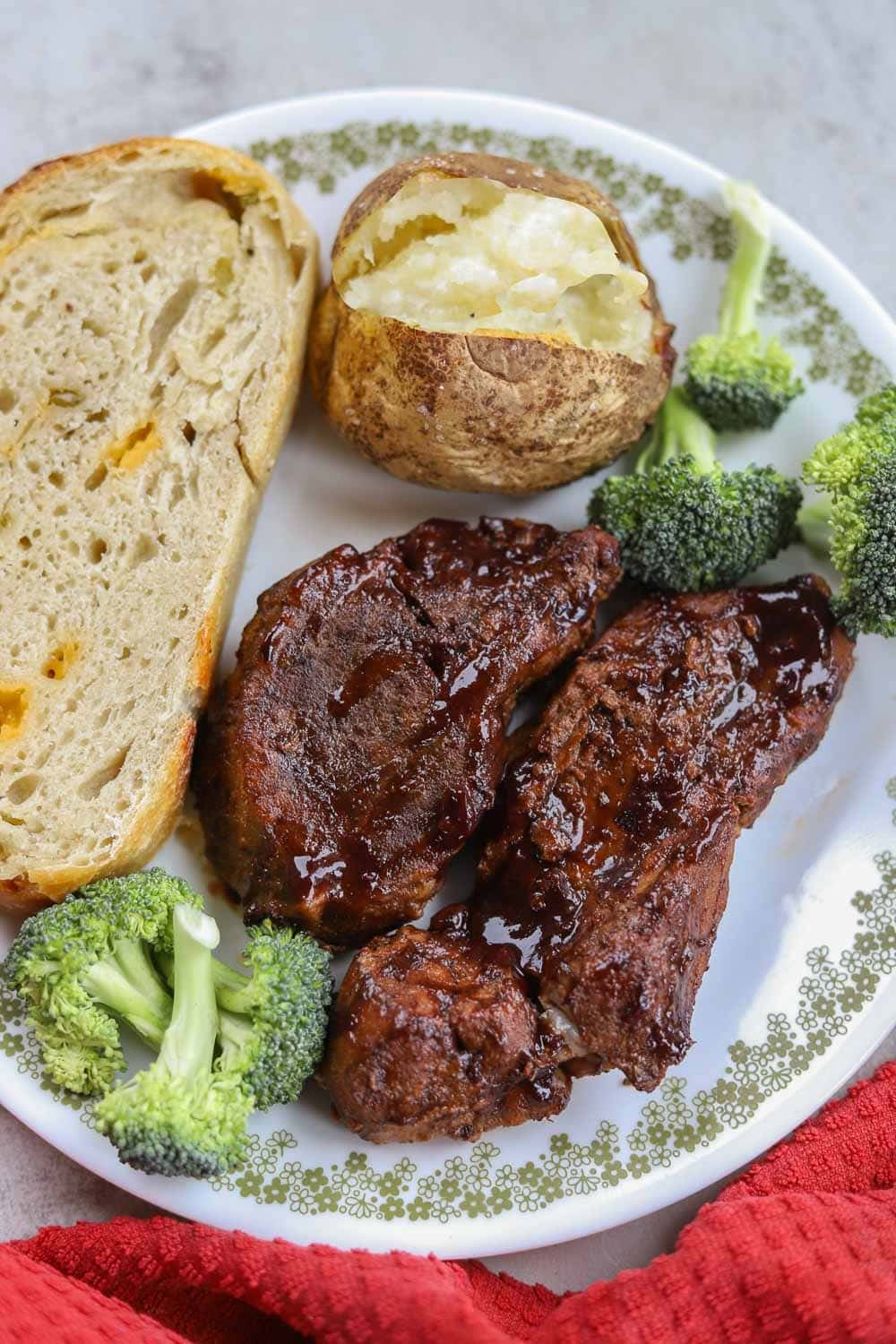 slow cooker country style ribs with baked potato, broccoli, and sourdough bread on a flowered white plate with a red napkin