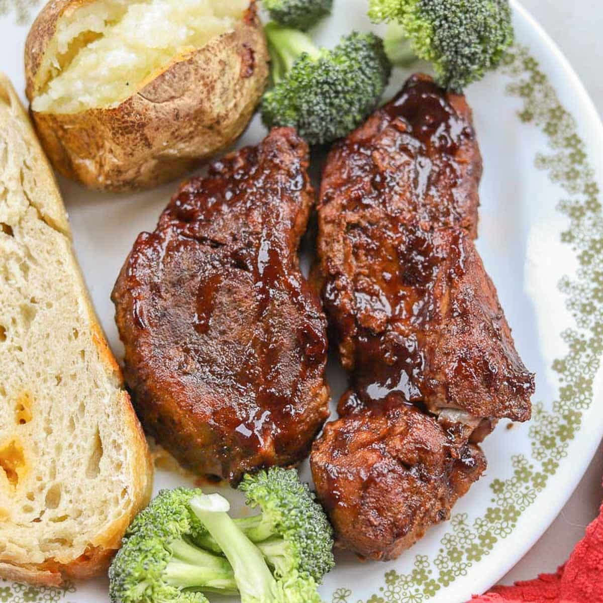 slow cooker country style ribs with baked potato, broccoli, and sourdough bread on a white plate next to a red napkin