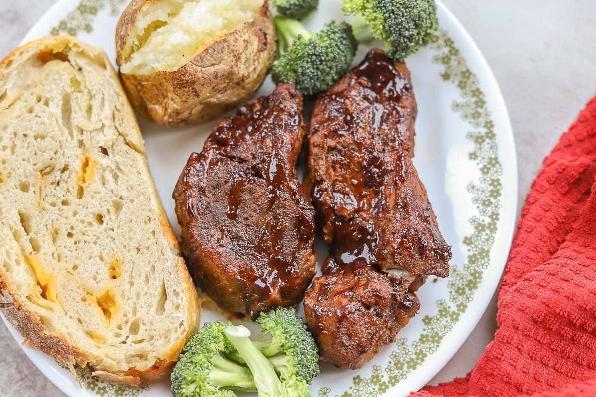 slow cooker country style ribs with baked potato, broccoli, and sourdough bread on a white plate next to a red napkin