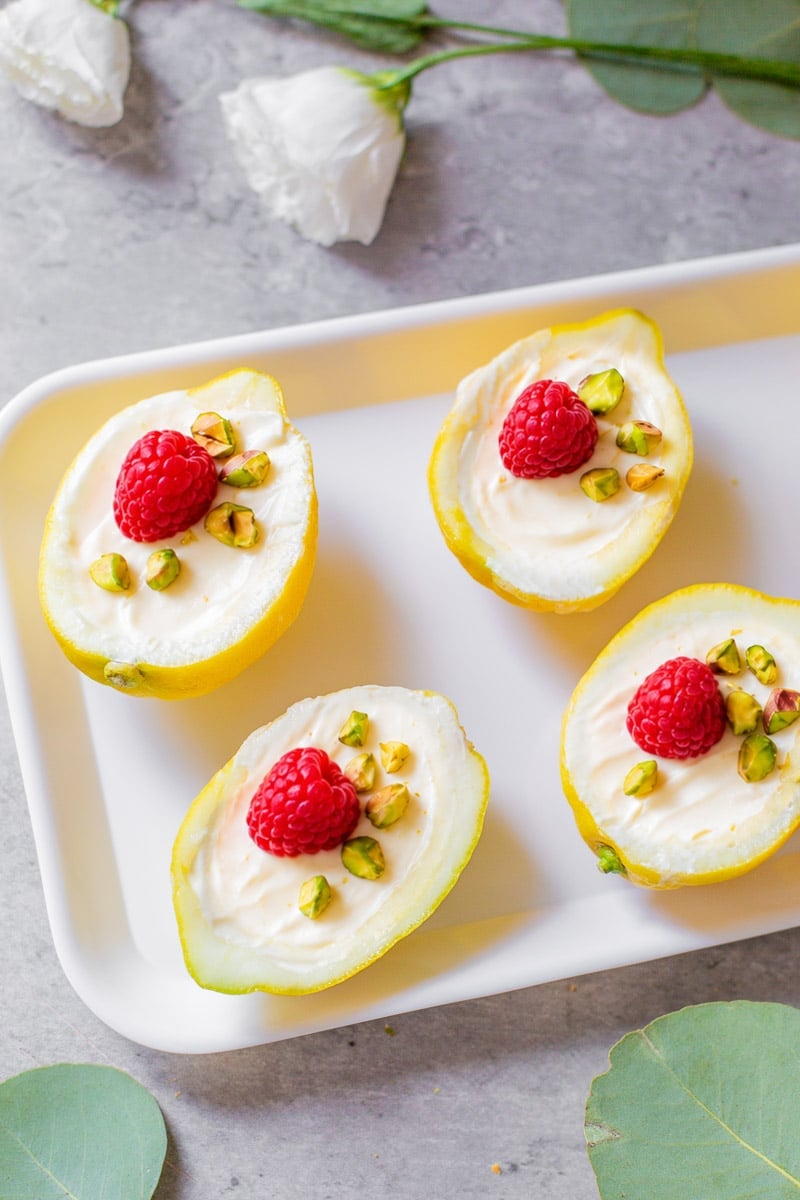 lemon posset in lemon skins with raspberries and pistachios beside eucalyptus and flowers.