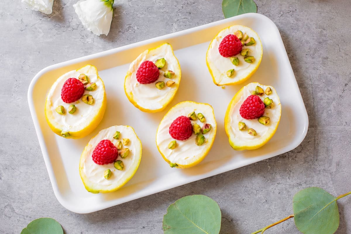lemon posset in lemon skins with raspberries and pistachios beside eucalyptus and flowers.