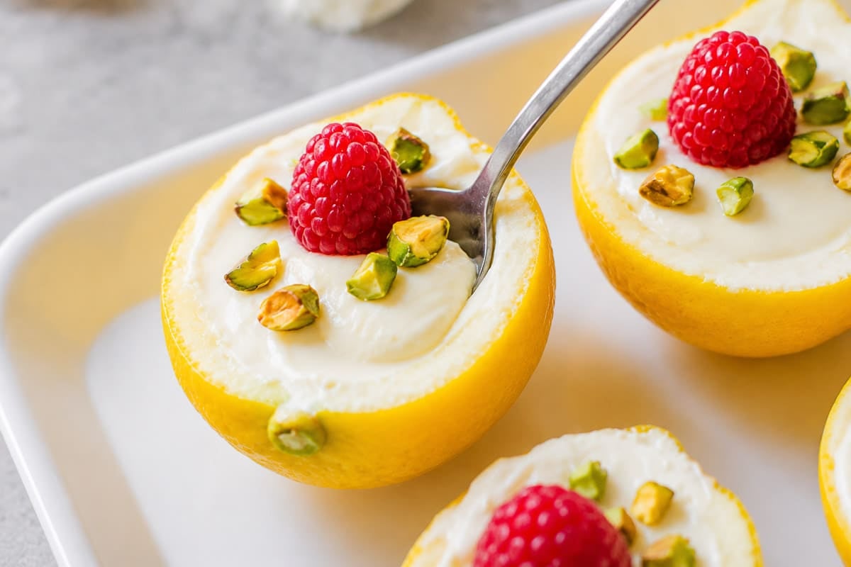 lemon posset in lemon skins with raspberries and pistachios and a spoon.