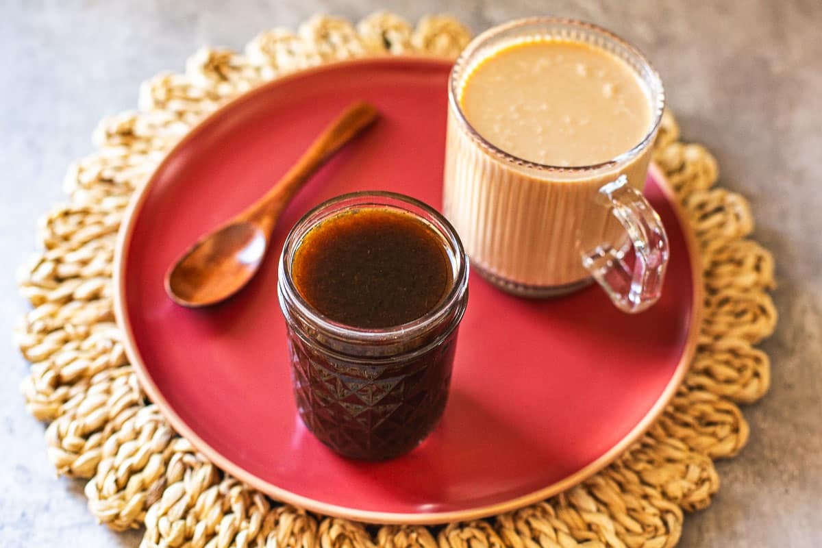 pumpkin spice syrup in jar beside cup of coffee and wooden spoon on a red plate over a straw mat.