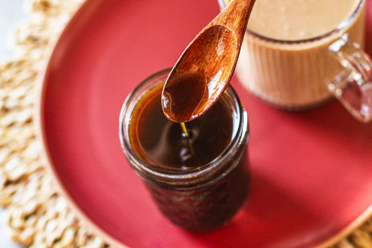pumpkin spice syrup drizzling from wooden spoon into jar beside cup of coffee.