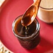 pumpkin spice syrup drizzling from wooden spoon into jar beside cup of coffee.
