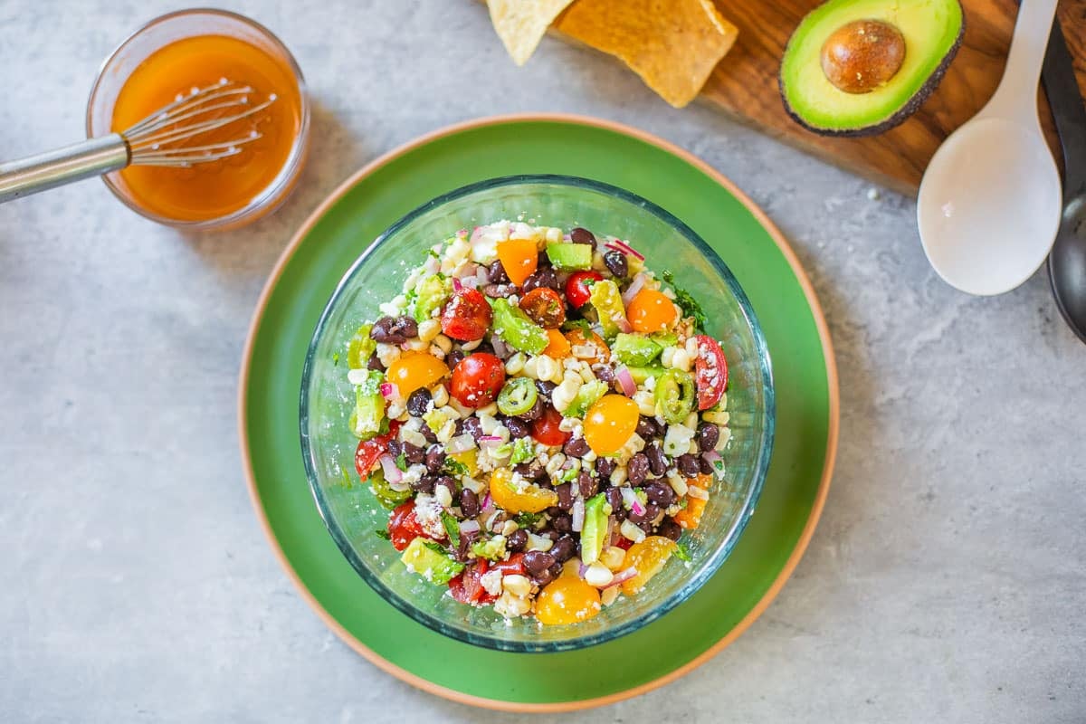 black bean and corn salsa beside vinaigrette, chips, avocado, salad tongs.