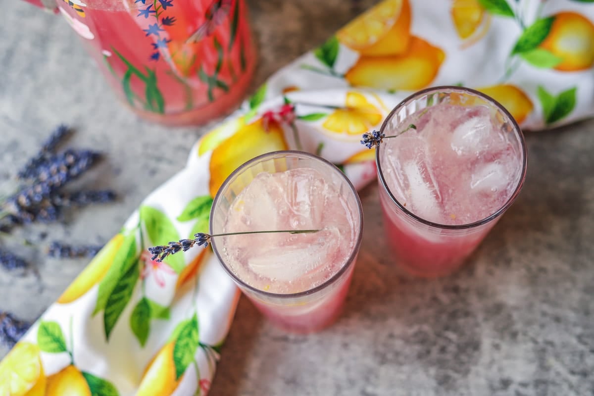 2 glasses of lavender lemonade with a pitcher full behind them and a towel with lemons and dried lavender next to them.