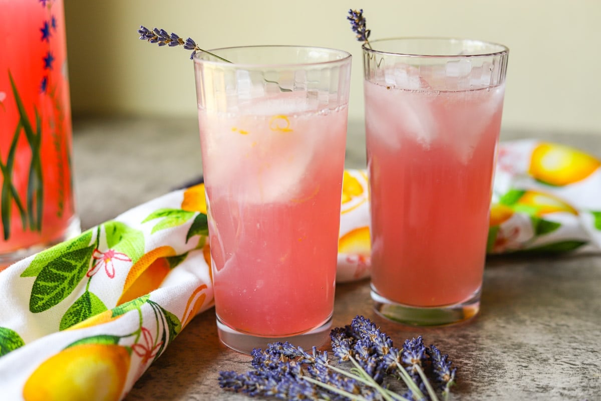 two glasses of lavender lemonade with ice with a dish towel with lemons behind them and lavender sprigs in front of them