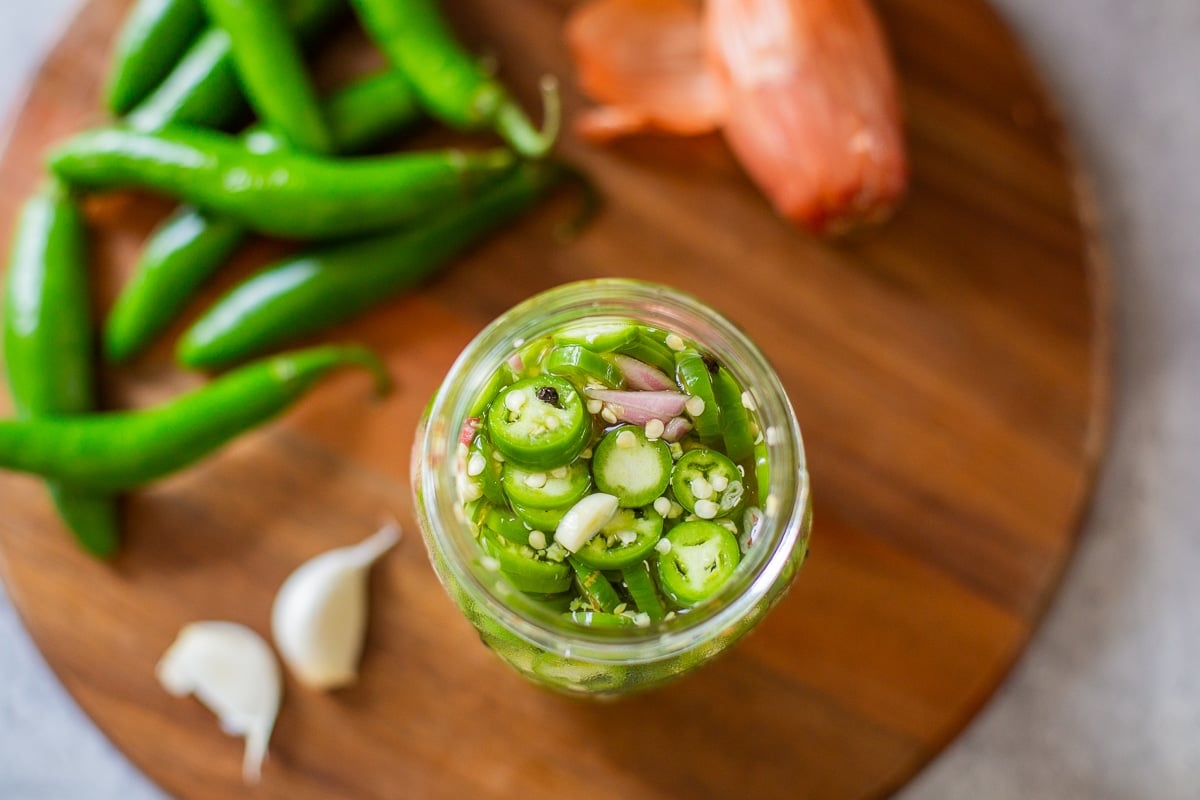 pickled serrano peppers in jar beside garlic, peppers, and shallots.
