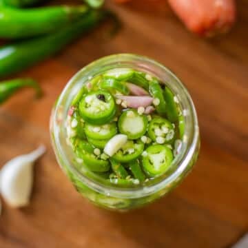 pickled serrano peppers in jar beside garlic, peppers, and shallots.