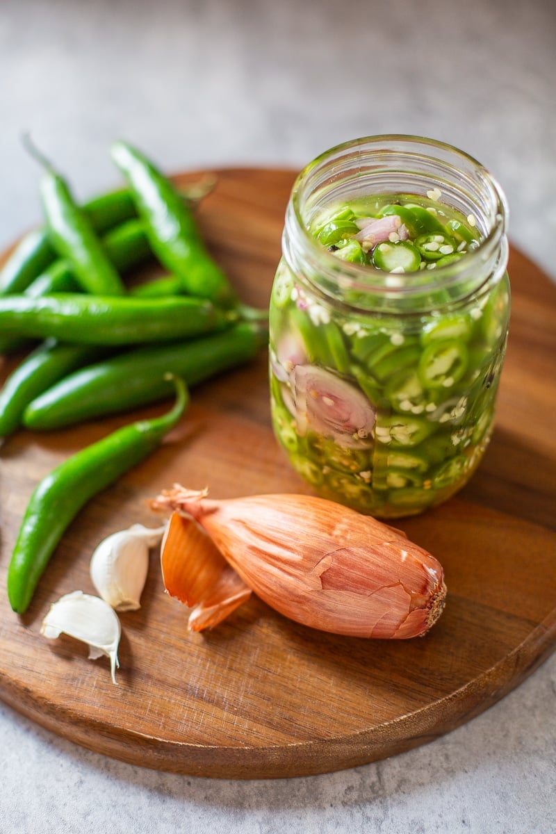 pickled serrano peppers in jar beside garlic, peppers, and shallots.